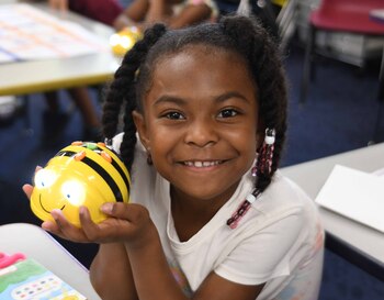 A young, black, female student holds a bumble bee technology device while smiling at the camera.