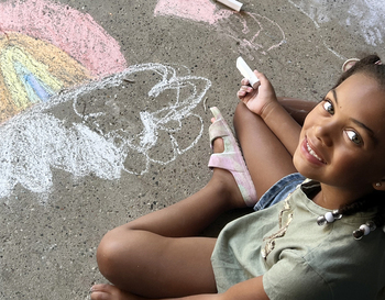 Young child sitting on the ground with a piece of sidewalk chalk in her hand. Ground has a rainbow and cloud drawn on it with sidewalk chalk.