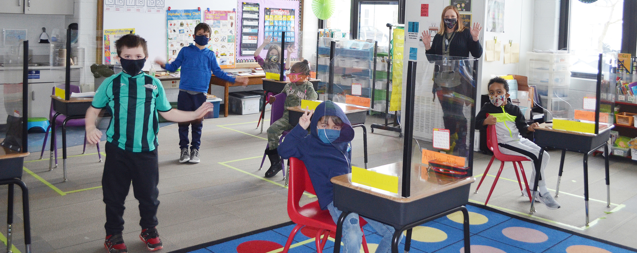 Children sitting at desks and standing in the classroom with face masks on, with their teacher.
